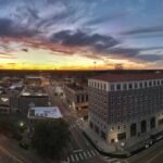 This image shows a panoramic view of downtown texarkana during a stunning sunset. The sky is painted with vibrant shades of orange, yellow, and blue as the sun sets on the horizon. The city's buildings are prominently visible, including historic structures and modern buildings. Streets intersect the downtown area, with a few vehicles parked along the sides. The city's architecture and layout are highlighted, creating a picturesque and serene urban scene as day turns to night.